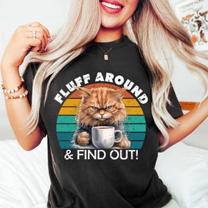 Könnte beinhalten: Schwarzes T-Shirt mit einem wütenden, orangefarbenen Kater, der eine Kaffeetasse hält. Der Text "FLUFF AROUND & FIND OUT!" ist über und unter dem Katzenbild vor einem Retro-Sonnenuntergangsdesign angeordnet.