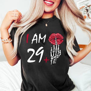 Könnte beinhalten: Schwarzes T-Shirt mit dem Text "I AM 29+". Das Design zeigt eine rote Glitzer-Lippen-Grafik, die den Mittelfinger zeigt, und eine weiße Skeletthand. Das Shirt hat einen Rundhalsausschnitt.
