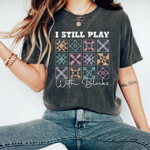May include: Dark gray t-shirt with the text "I Still Play With Blocks." The shirt features a grid of colorful quilt block designs in shades of orange, blue, pink, and purple. The shirt is a comfortable, casual style.