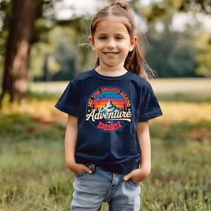Peut inclure: Un t-shirt bleu marine avec l'inscription "Let the Second Grade Adventure Begin" dans un cercle. Le graphisme comprend une chaîne de montagnes, des oiseaux et un arc-en-ciel. Le t-shirt est porté par une jeune personne dans un cadre extérieur herbeux.