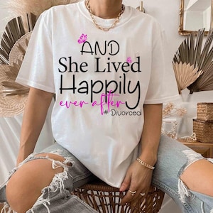 Könnte beinhalten: Weißes T-Shirt mit schwarzem Text, der "AND She Lived Happily ever after #Divorced" lautet. Der Text ist mit rosa Schmetterlingsgrafiken verziert.