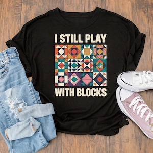 May include: Black t-shirt with the text "I STILL PLAY WITH BLOCKS" above a colorful quilt block design. The shirt is paired with blue jeans and white and pink sneakers on a wooden surface.