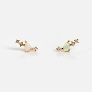 May include: A pair of gold stud earrings, each featuring a central pear-shaped opal gemstone flanked by two small, clear, round-cut stones on either side, arranged in a delicate, asymmetrical, curved cluster design on a gold metal base.