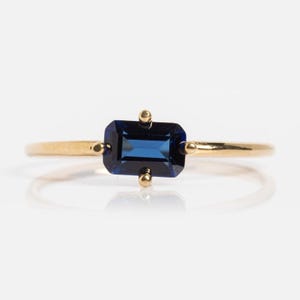 Op de afbeelding: Een gouden ring met een rechthoekige, diepblauwe edelsteen. De edelsteen is gezet in een gouden band met kleine gouden accenten aan weerszijden. De ring wordt gepresenteerd op een witte achtergrond, wat het elegante ontwerp benadrukt.