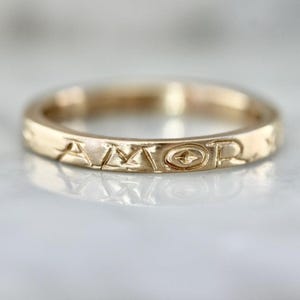 May include: A gold-colored ring with the word "AMOR" engraved around the band. The ring has a simple, elegant design, with the letters carved in a classic font. The ring is likely made of gold and has a polished finish.