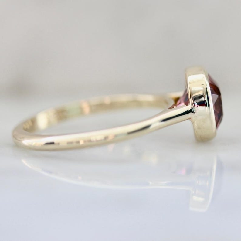 Hot Honey Pink-Peach Tourmaline Ring featuring a 1.81 carat cushion cut lab-created tourmaline set in 14k yellow gold. The gemstone glows with vibrant hues of pink, peach, orange, and red that shift in different lighting. A rounded bezel setting keeps the design sleek and modern, while the slim 1.71 mm band makes it perfect for stacking with other rings. A versatile and radiant gemstone ring that blends everyday luxury with a bold pop of color.