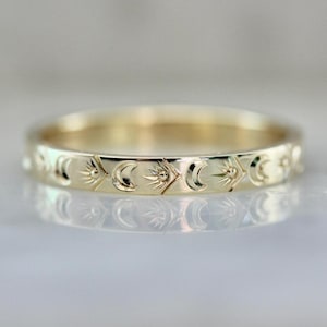 May include: A gold-colored ring with a repeating pattern of sun and crescent moon designs. The ring is highly polished and reflects light, creating a bright and shiny appearance. The ring is a delicate piece of jewelry.
