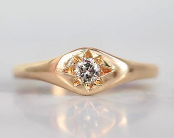 Könnte beinhalten: Ein goldener Ring mit einem klaren Edelstein, der in einem achtzackigen Sternmuster gefasst ist. Der Ring hat einen glatten, polierten Ring und eine erhöhte Fassung für den Edelstein. Der Ring wird auf einer weißen Oberfläche präsentiert.