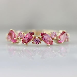 Puede incluir: Un anillo de oro con un grupo de piedras preciosas rosas. El anillo tiene un diseño delicado con piedras de talla marquesa y redondas. Las piedras preciosas son de un color rosa vibrante, engastadas en la banda de oro.