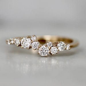 Könnte beinhalten: Ein goldener Ring mit einer Ansammlung funkelnder, rund geschliffener Diamanten. Die Diamanten sind in einem zarten, asymmetrischen Muster angeordnet, wodurch ein einzigartiges und elegantes Design entsteht. Der Ring hat ein glattes, poliertes Goldband.