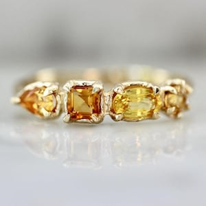 May include: A gold ring with a sequence of yellow gemstones in different shapes. The gemstones include a pear-shaped, a square-cut, and an oval-cut stone. The ring is set in a gold band.