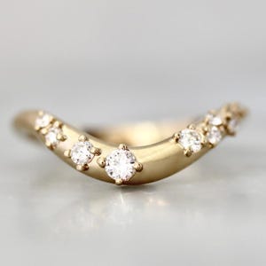 May include: A curved gold ring with several small, sparkling diamonds set along the top. The ring has a smooth, matte finish and a delicate, elegant design. The diamonds are evenly spaced and catch the light, creating a subtle sparkle.