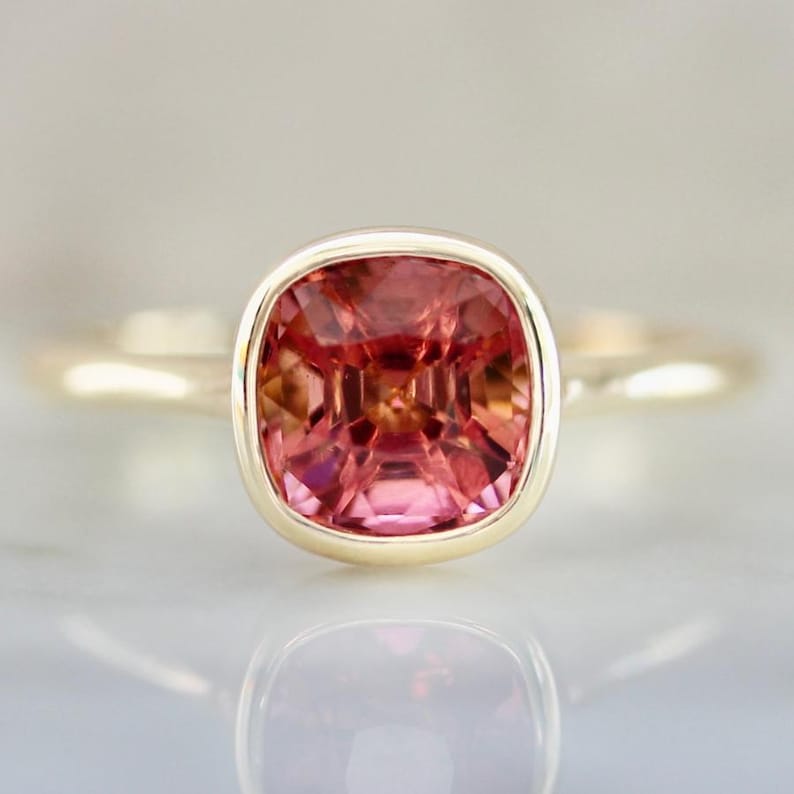 Hot Honey Pink-Peach Tourmaline Ring featuring a 1.81 carat cushion cut lab-created tourmaline set in 14k yellow gold. The gemstone glows with vibrant hues of pink, peach, orange, and red that shift in different lighting. A rounded bezel setting keeps the design sleek and modern, while the slim 1.71 mm band makes it perfect for stacking with other rings. A versatile and radiant gemstone ring that blends everyday luxury with a bold pop of color.