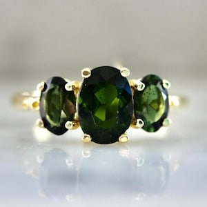 Pode incluir: Um anel de ouro com três pedras preciosas ovais verde-escuras. A pedra central é a maior, ladeada por duas pedras menores. A banda e as engastes do anel são de ouro, criando um design clássico.