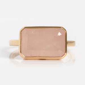 May include: A gold ring featuring a large, rectangular, light pink gemstone. The gemstone is set within a gold bezel, and the band is a simple, thin gold design. The ring is displayed on a white background.