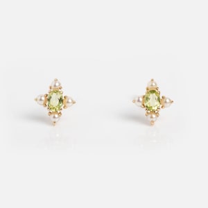 May include: A pair of gold stud earrings. Each earring has an oval peridot gemstone at the centre, encircled by four small pearls. The earrings are in a cross shape. The background is white.