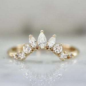 Puede incluir: Un anillo dorado con un diseño en forma de corona. El anillo presenta varias piedras preciosas claras y brillantes, incluyendo piedras en forma de pera y redondas. El anillo está sobre una superficie reflectante.