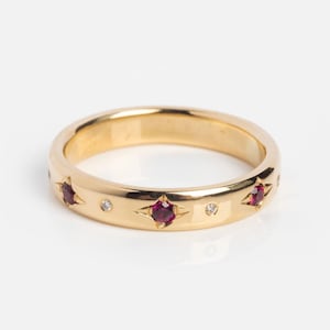 May include: A gold ring with a polished finish. The ring features star-shaped settings with red gemstones and small, round, clear gemstones. The ring is set against a white background.