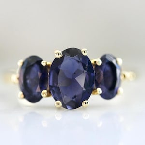 Puede incluir: Un anillo de oro con tres piedras preciosas ovales de color azul oscuro. Las piedras están dispuestas en fila, con una banda y garras doradas. El anillo se muestra sobre una superficie blanca.