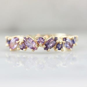 May include: A gold ring featuring a cluster of purple gemstones in various shapes and shades. The gemstones are set closely together, creating a floral or organic design. The ring's band is a simple, polished gold.