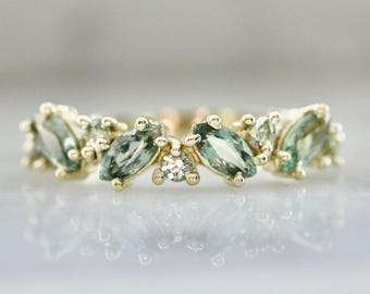 May include: A gold ring featuring a cluster of light green marquise-cut gemstones and small round accent stones. The ring's design is symmetrical, with the gemstones arranged in a floral or leaf-like pattern. The ring is set against a reflective surface.