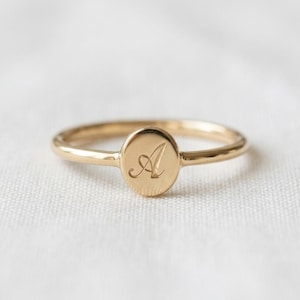 May include: A gold ring featuring a polished, oval-shaped face engraved with a cursive letter A. The band is thin and smooth, maintaining a consistent metallic finish throughout the entire piece, showcasing a simple and refined design.