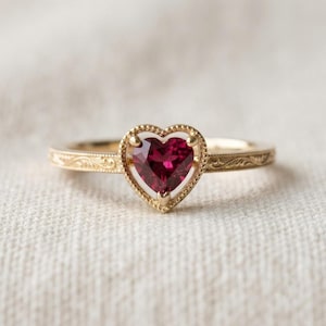 May include: A gold ring featuring a heart-shaped deep red gemstone set within a beaded frame. The band of the ring displays an intricate engraved pattern along its surface, extending from the central heart setting toward the sides of the band.
