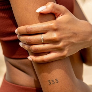 May include: A delicate gold ring featuring a floral design with small, sparkling gemstones. The ring is worn on a finger, showcasing its intricate details. The number "333" is tattooed on the arm. The ring is a piece of jewelry.