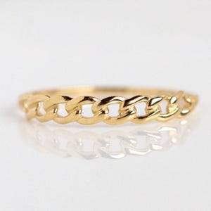 May include: A gold-colored chain ring. The ring features a continuous chain link design. The ring is displayed on a reflective surface, which mirrors the ring's design. The ring is a simple, elegant piece of jewelry.