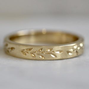 May include: A gold ring with a leaf pattern. The ring is a band style with a matte finish. The leaves are etched into the surface of the ring. The ring is a warm gold color.