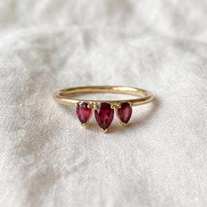 May include: A gold ring featuring three pear-shaped red gemstones set in a row. The central stone is slightly larger than the two flanking stones, all secured by small prongs on a thin, polished gold band with a smooth, rounded finish.