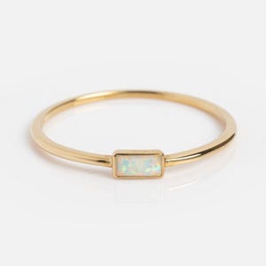 Puede incluir: Un anillo de oro con una piedra de ópalo rectangular. El anillo tiene una banda delgada y un diseño simple y elegante. La piedra de ópalo tiene una apariencia blanca e iridiscente.
