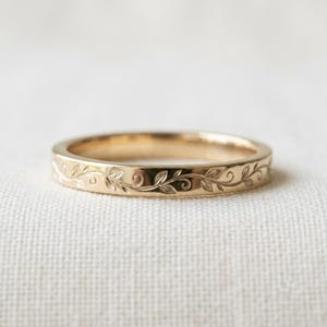 Puede incluir: Un anillo de banda de oro con un patrón grabado continuo de delicadas enredaderas sinuosas y pequeñas hojas que rodean toda la superficie exterior del metal. El anillo tiene un acabado pulido y un perfil delgado y uniforme en todo su diseño.