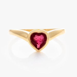 May include: A gold ring with a heart-shaped, deep red gemstone. The ring has a smooth, polished band that curves up to hold the heart-shaped stone. The gemstone is set in a bezel setting, enhancing its shape and colour.