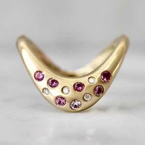 May include: A gold-colored ring with a curved design, adorned with a series of pink gemstones and small, clear diamonds. The ring's smooth surface reflects light, highlighting the contrast between the gold and the colorful stones.
