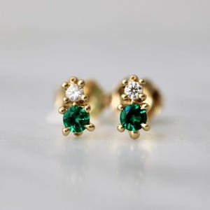 May include: A pair of gold stud earrings, each featuring a small, clear, round-cut stone positioned above a larger, round-cut emerald green stone. The stones are held in place by gold prongs, creating a vertical, two-stone design on each earring.