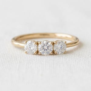 May include: A gold ring featuring three round, clear, faceted stones set in a horizontal row. The band is smooth and polished, with each stone secured by four small prongs. The design highlights the brilliance of the stones against the gold metal.
