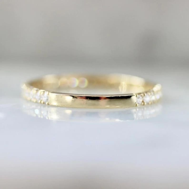 Puede incluir: Un anillo de banda de oro con una fila de peque&ntilde;as piedras preciosas transparentes engastadas en la parte superior y una banda de oro lisa y pulida. El anillo est&aacute; sobre un fondo suave y borroso.