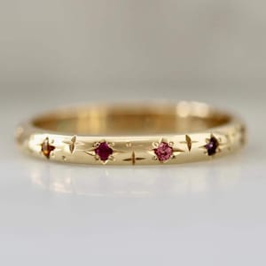 May include: A gold ring featuring star and cross patterns. The ring is set with small gemstones in shades of red and pink. The ring has a polished finish and a classic design, suitable for everyday wear or special occasions.