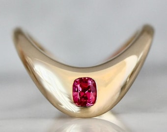 May include: A gold-colored ring with a curved design, featuring a vibrant, cushion-cut, red gemstone. The ring's polished surface reflects light, highlighting the gemstone's color and shape. The ring is set on a reflective surface.