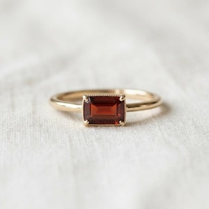 May include: A gold ring featuring a rectangular, emerald-cut deep red garnet gemstone set in a four-prong setting with a delicate beaded border detail around the stone, mounted on a smooth, thin, polished gold band.