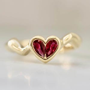 May include: A gold ring with a heart-shaped setting, containing two red gemstones. The ring has a smooth, polished band that curves around the heart-shaped centrepiece. The gemstones are a deep red colour, set in a gold bezel.
