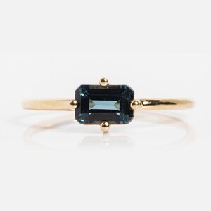 Op de afbeelding: Een gouden ring met een rechthoekige, donkerblauwe edelsteen. De edelsteen is gezet in een gouden band met kleine gouden accenten. De ring wordt gepresenteerd tegen een witte achtergrond, wat het elegante ontwerp benadrukt.