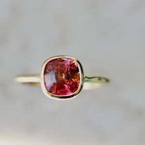 Hot Honey Pink-Peach Tourmaline Ring featuring a 1.81 carat cushion cut lab-created tourmaline set in 14k yellow gold. The gemstone glows with vibrant hues of pink, peach, orange, and red that shift in different lighting. A rounded bezel setting keeps the design sleek and modern, while the slim 1.71 mm band makes it perfect for stacking with other rings. A versatile and radiant gemstone ring that blends everyday luxury with a bold pop of color.