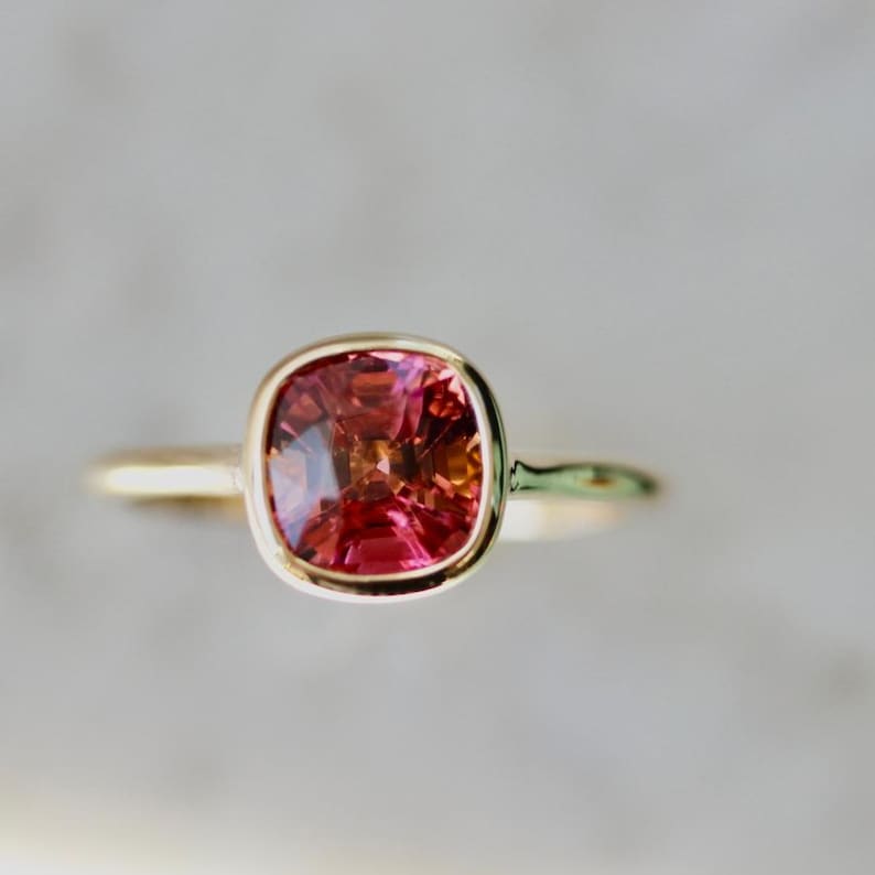 Hot Honey Pink-Peach Tourmaline Ring featuring a 1.81 carat cushion cut lab-created tourmaline set in 14k yellow gold. The gemstone glows with vibrant hues of pink, peach, orange, and red that shift in different lighting. A rounded bezel setting keeps the design sleek and modern, while the slim 1.71 mm band makes it perfect for stacking with other rings. A versatile and radiant gemstone ring that blends everyday luxury with a bold pop of color.