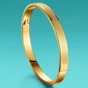 May include: A gold-coloured bangle bracelet with a textured pattern on the top surface. The bracelet has a hinged clasp and a smooth, polished finish on the sides. The background is a solid teal colour.
