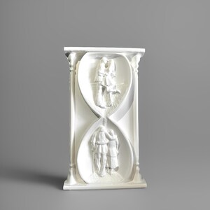Puede incluir: Escultura blanca rectangular con forma de reloj de arena. La escultura presenta tallas en bajorrelieve de una pareja abrazándose y besándose. El diseño es de temática romántica, adecuado para la decoración del hogar.