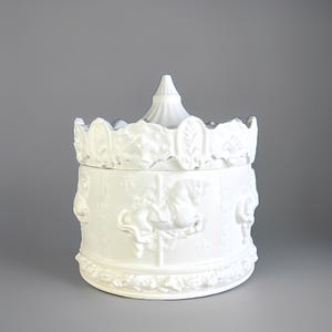 May include: White ceramic jar with a carousel design. The jar has embossed carousel horses and floral details. The lid has a conical top and a scalloped edge. A decorative storage container.