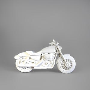 Puede incluir: Modelo de motocicleta blanca con un diseño detallado, que incluye ruedas, asiento y manillar. La motocicleta se encuentra sobre un fondo gris, mostrando su forma y características. El modelo parece ser una réplica en miniatura.