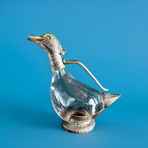 May include: A clear glass duck-shaped decanter with a silver-coloured metal head, neck, handle, and base. The duck's head is detailed with textured scales and a golden beak. The decanter sits on a round, textured base against a blue background.